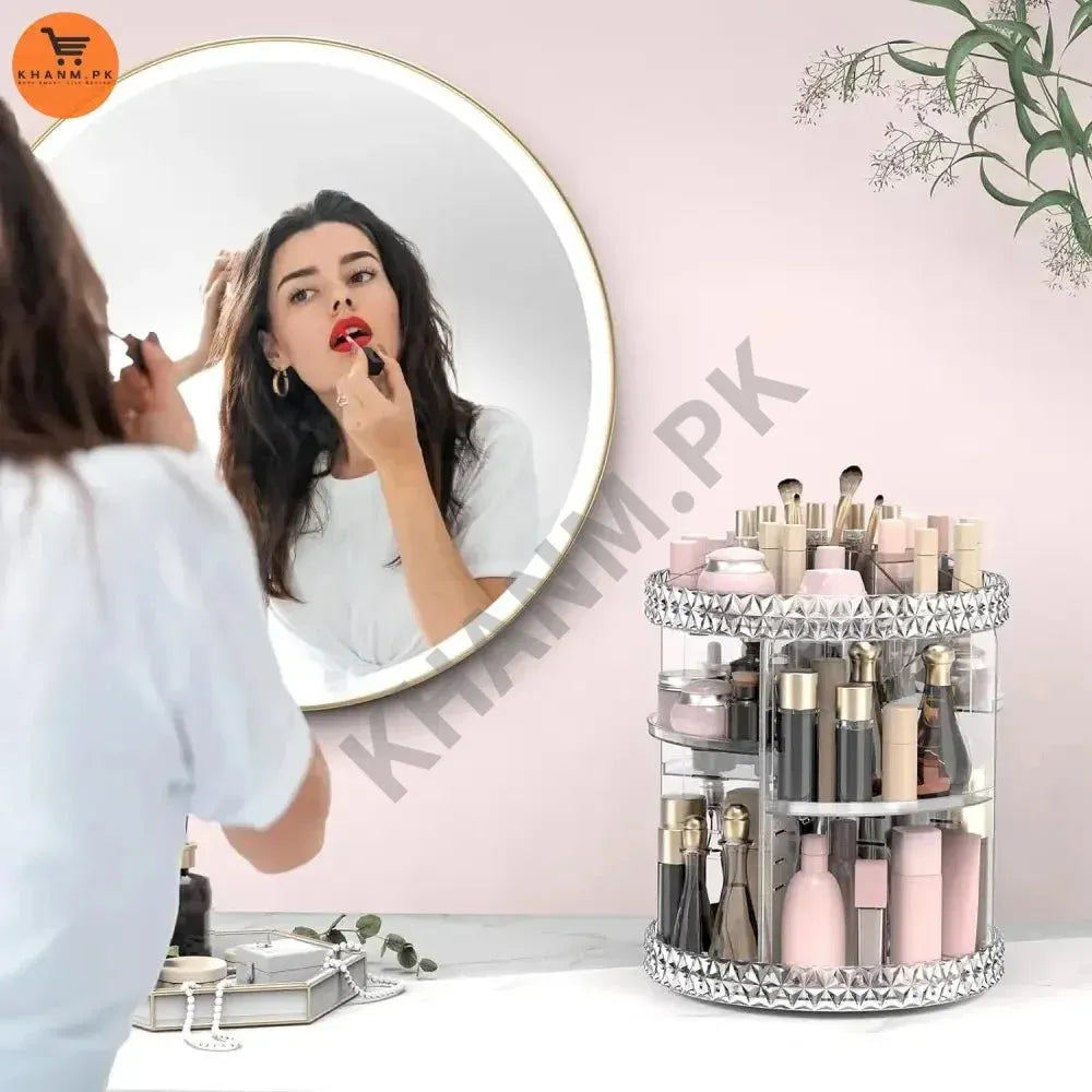 Woman applying makeup in front of a mirror with a clear cylindrical makeup organizer filled with products on a pink wall background.
